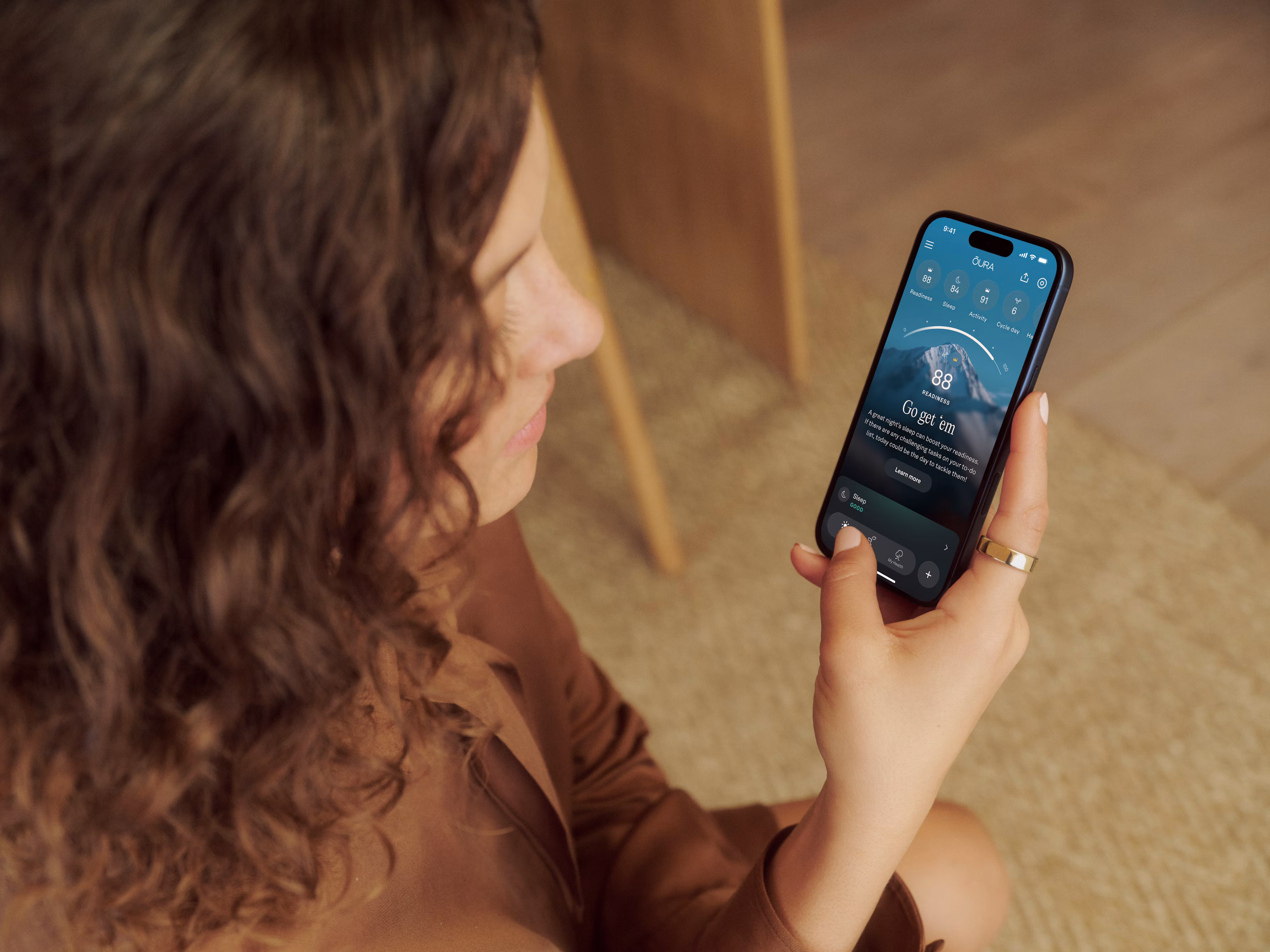 A woman with curly hair holding a smartphone, viewing the Oura app while wearing an Oura Ring 5.