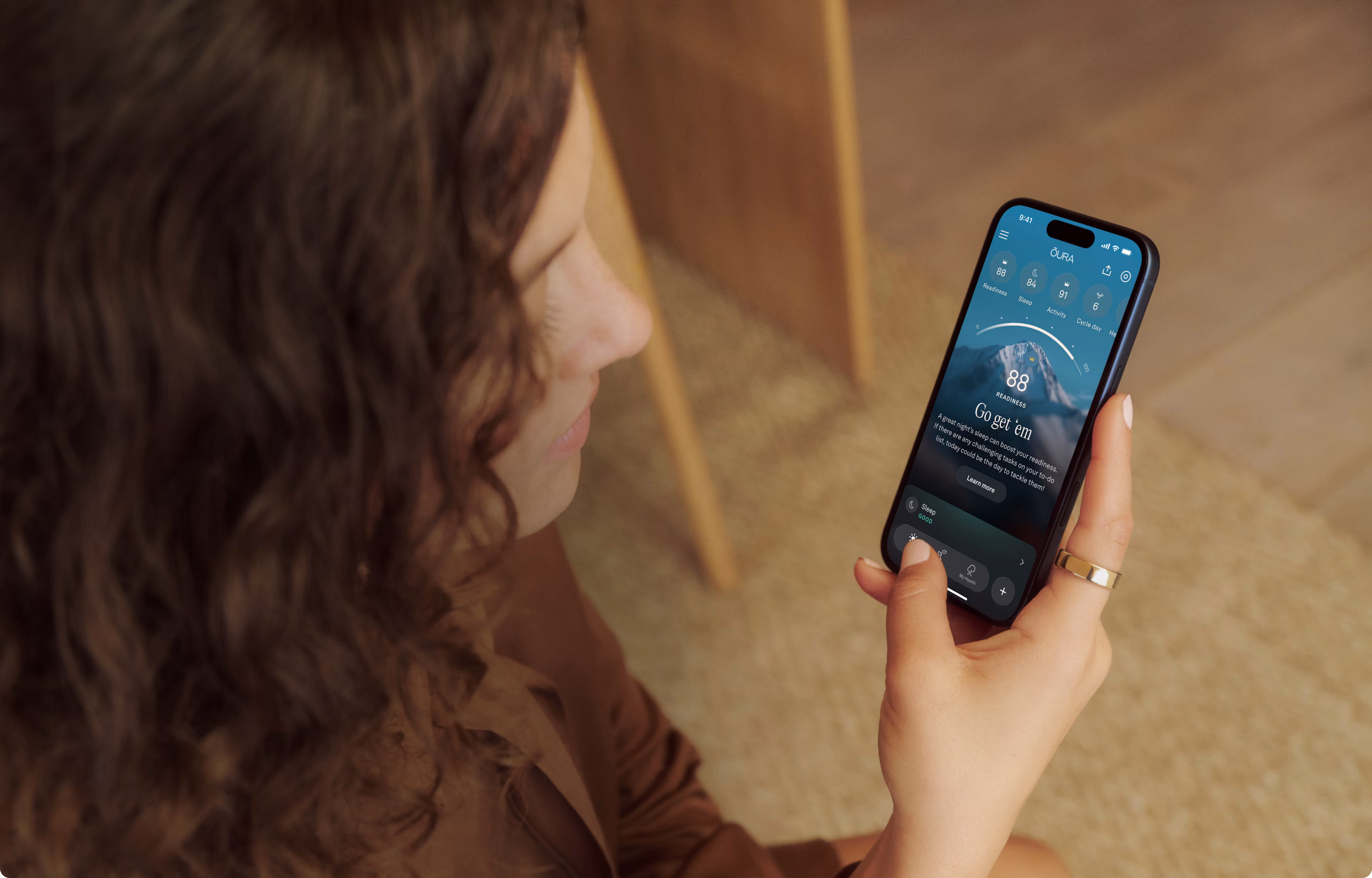 Woman with curly hair reviewing her Oura app readiness score on a smartphone, wearing a Gold Oura Ring