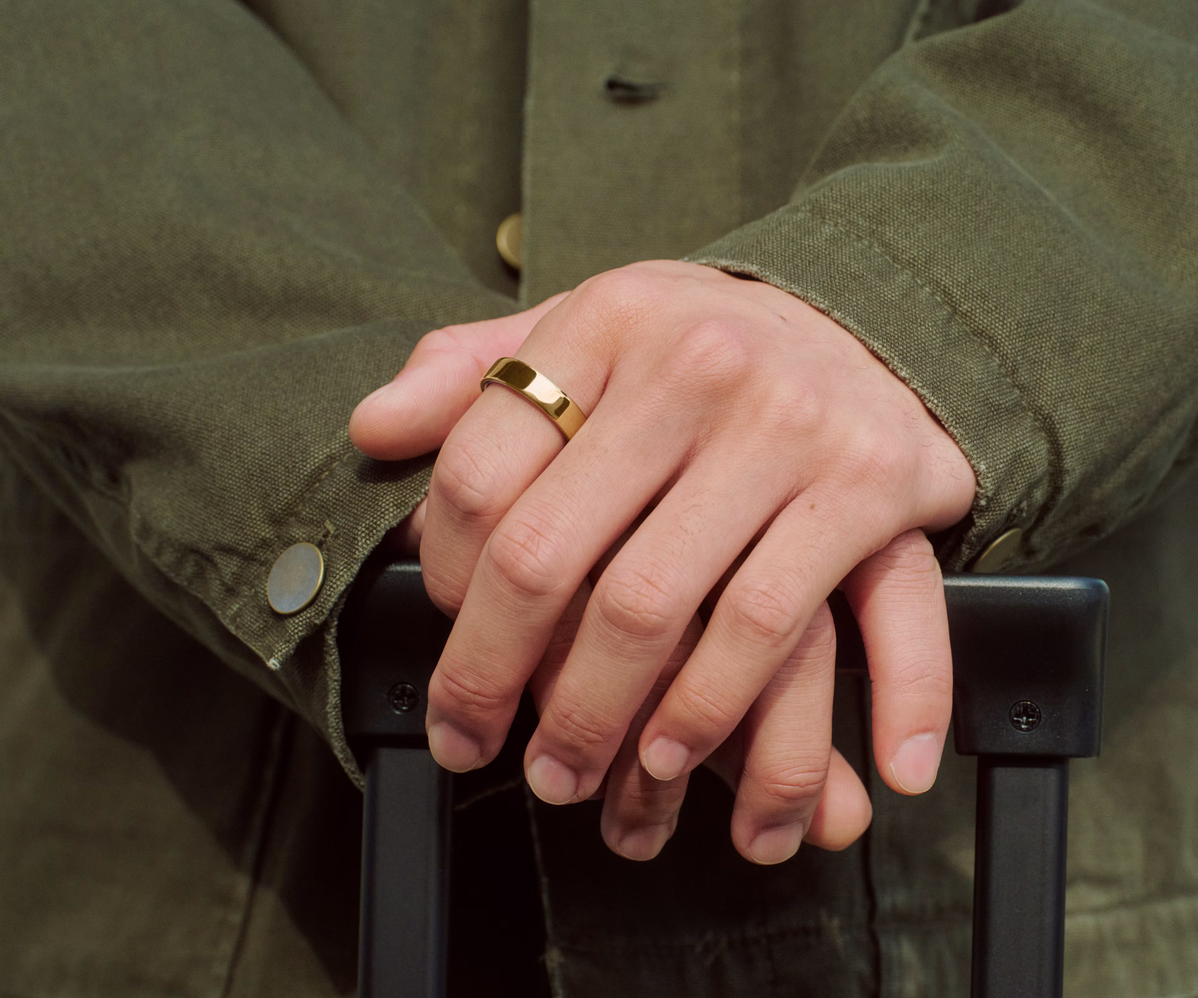Close up of a person's hands resting on the handle of a suitcase with a Gold Oura Ring 5 on their finger