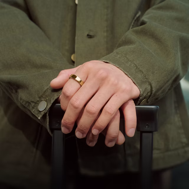 Close up of a person's hands resting on the handle of a suitcase with a Gold Oura Ring 5 on their finger