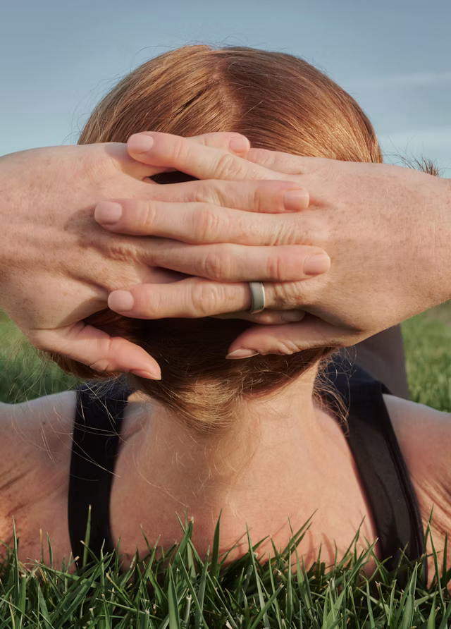 A person laying in grass with their hands cradling the back of their head while wearing an Oura Ring 5 in Brushed Silver finish