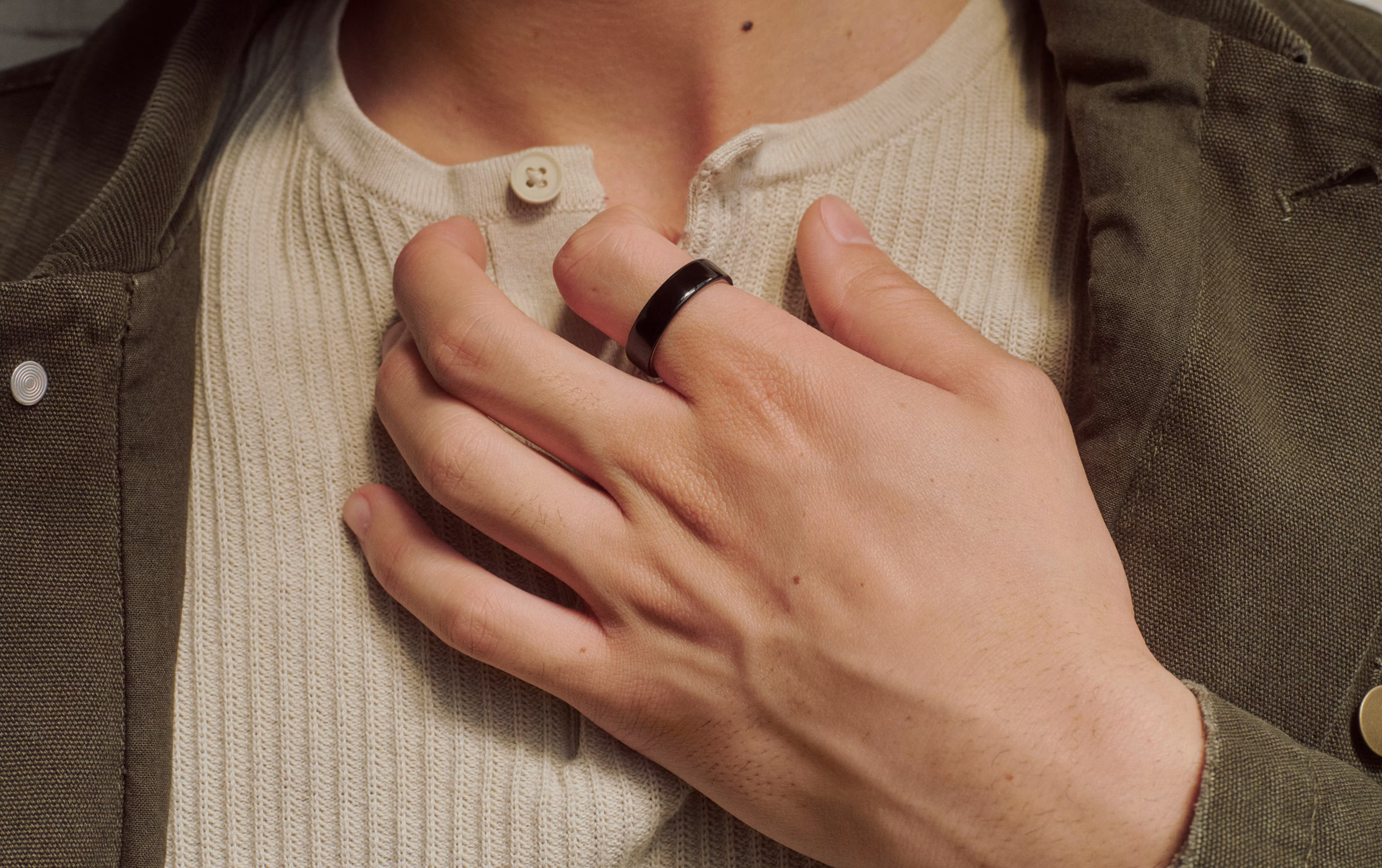Close-up of a hand wearing a Black Oura Ring placed over the chest with a cream henley and olive jacket.