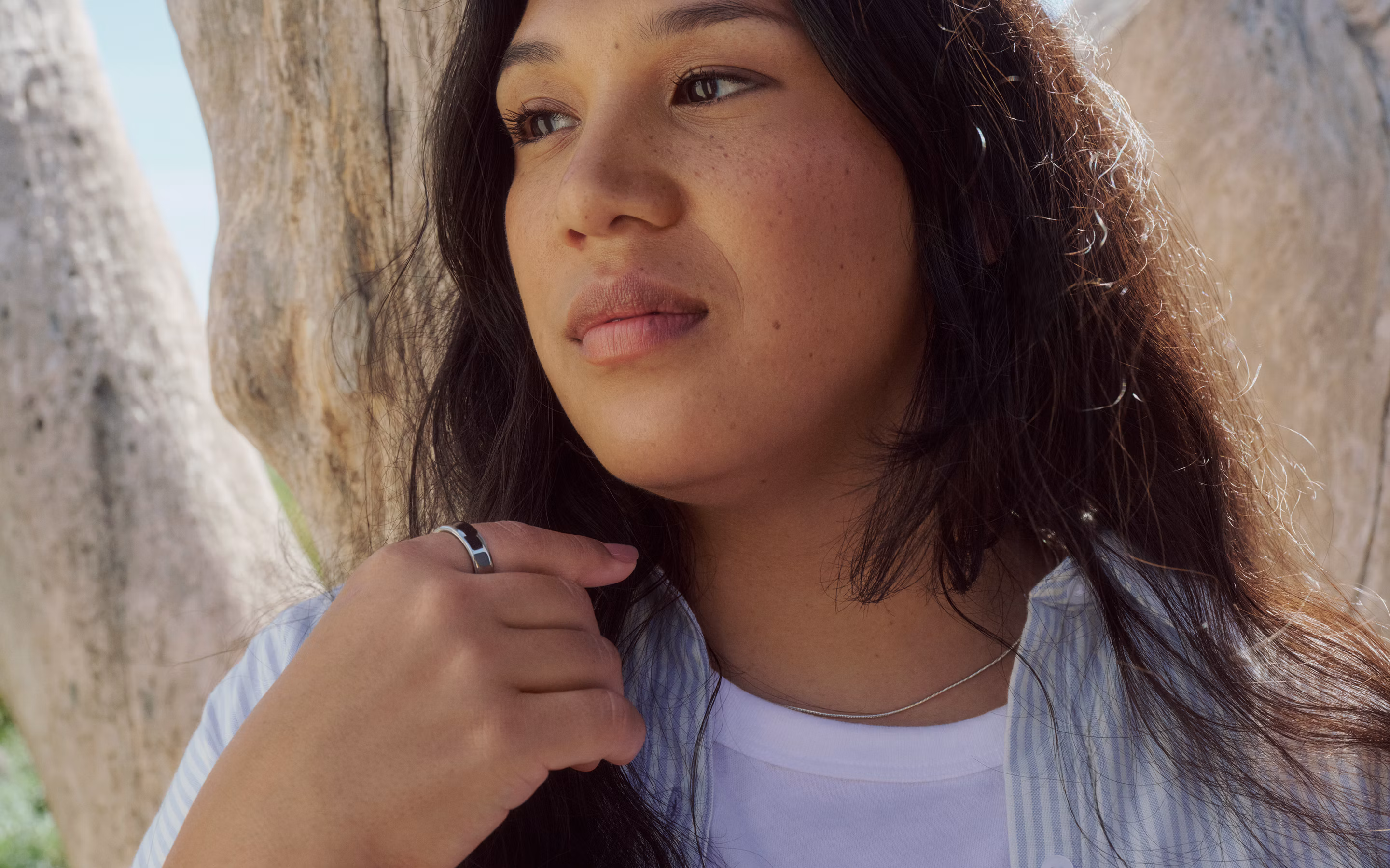 Woman outdoors near boulders wearing a Silver Oura Ring, hand touching her white shirt collar.