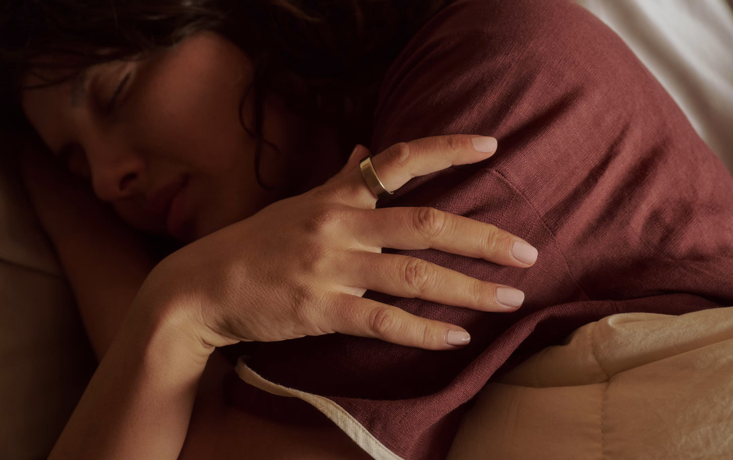 Woman sleeping peacefully with her hand draped over a mauve pillow, wearing a Gold Oura Ring.