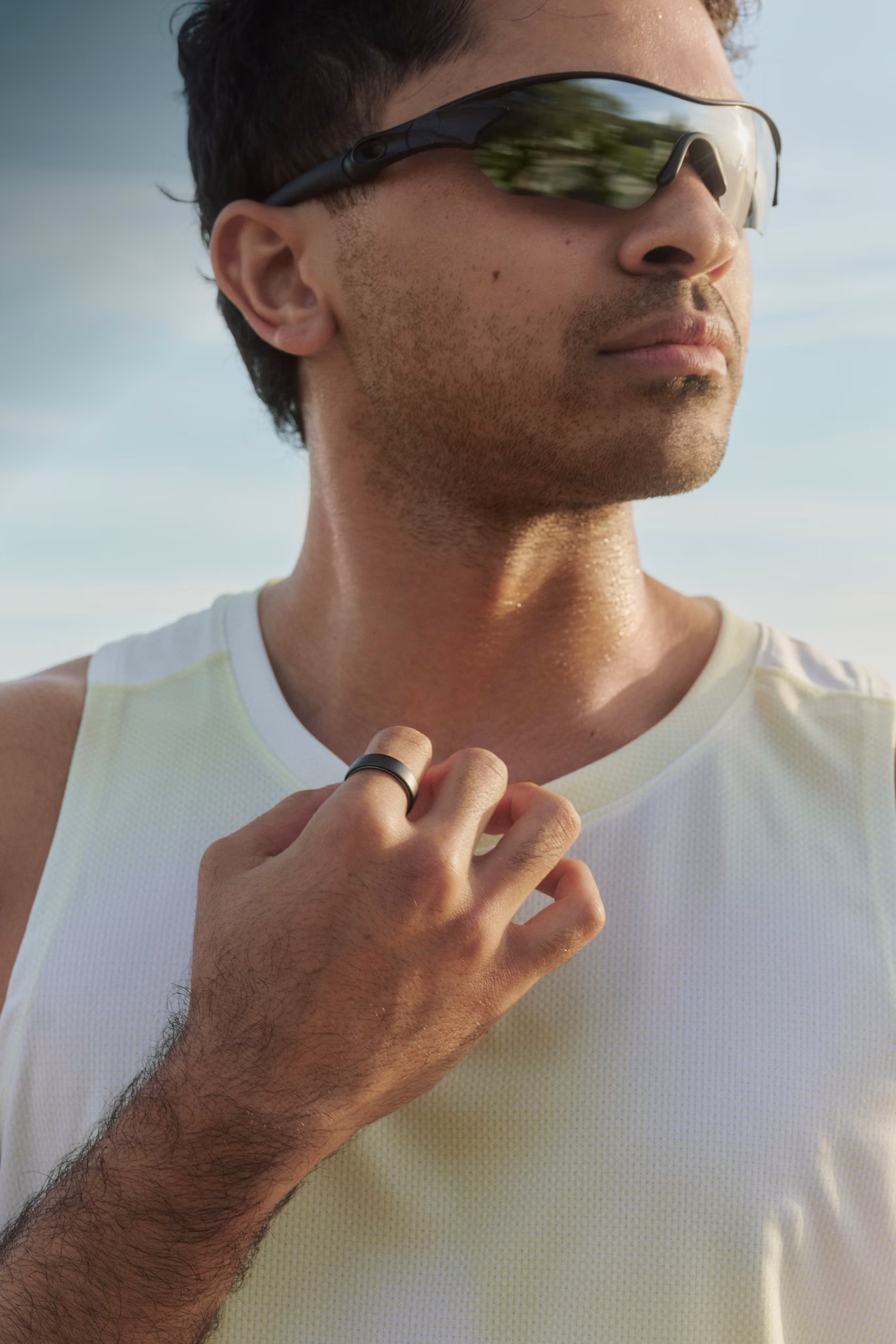 Man in sunglasses and white tank top wearing a Black Oura Ring, hand near his collar outdoors.