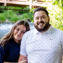 A woman wearing a silver oura ring and her partner smiling poses outdoors near a flowing river and wooden bridge surrounded by greenery.